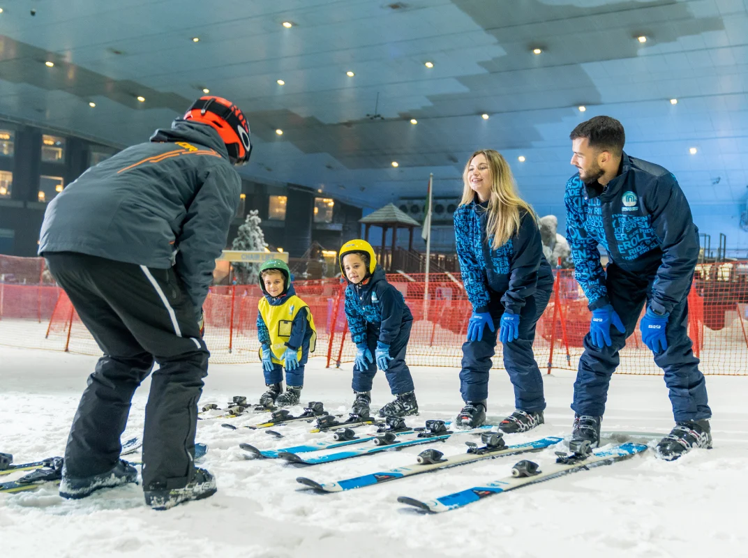 Children learning to ski indoors, guided by instructors, surrounded by snow and ski equipment in a vibrant ski facility.